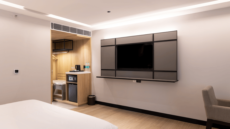 A Premium Room at Sands Point Hotel, Pondicherry, featuring a television mounted with a floating shelf below, a smart room control panel, and a grey upholstered armchair to the right.