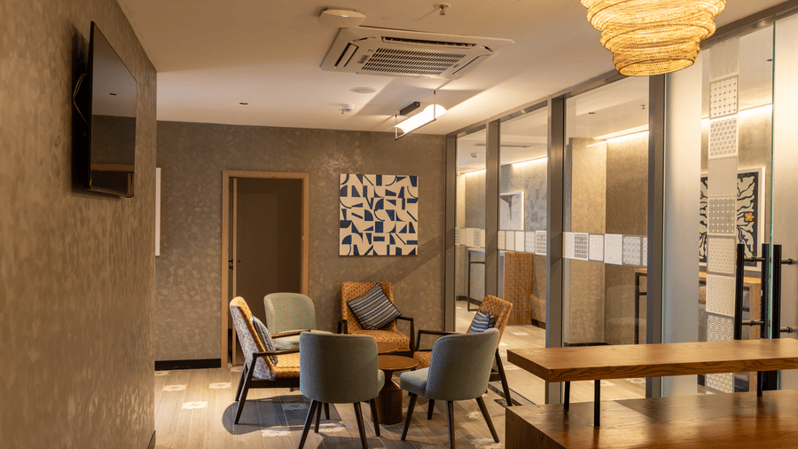 A serene and warmly lit spa waiting lounge at Sands Point Hotel featuring a cozy seating arrangement of sage green and orange upholstered armchairs with striped cushions around a small round wooden side table, a blue and beige geometric abstract artwork.