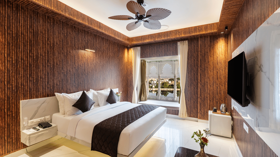 Panoramic view of a luxury hotel suite, showing the bed, a ceiling fan, textured timber walls, and bright natural lighting.