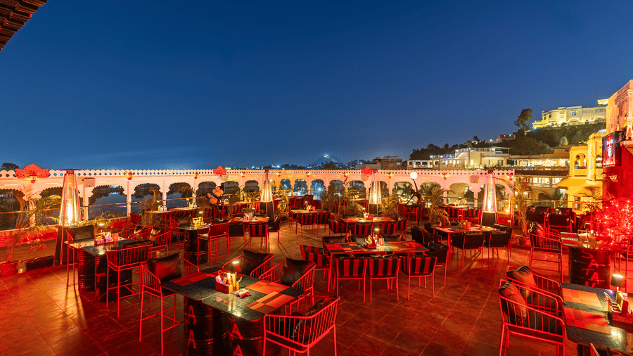 Open-air rooftop restaurant at night, featuring dark tables and chairs with a vibrant red glow and arched colonnade.