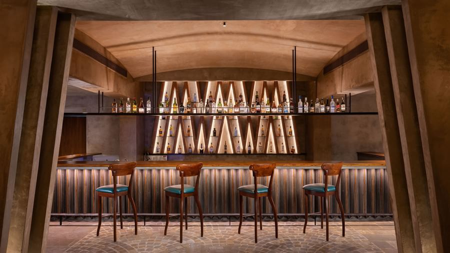 A dimly lit bar with a long counter, several bar stools, and shelves of bottles illuminated from behind, at Shilton International, Kochi.