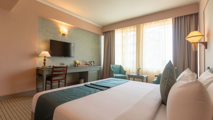  A guest room at Shilton International, Kochi, with a large bed, a desk area with a television, and a window with curtains.