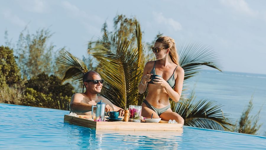 View of a man and woman with a Floating Breakfast platter in Infinity pool at Shooting Star boutique hotel. Ocean and trees can be found in the background.