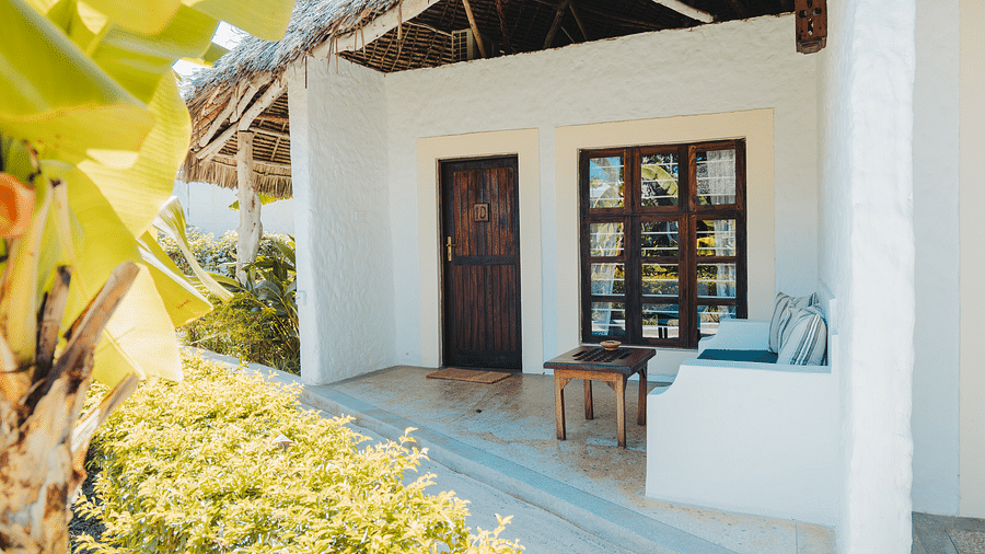 A bright, sunlit patio with a white wall, a dark wooden door, a multi-pane window, and a wooden table. Green foliage is visible in the foreground - shooting star Boutique Hotel.