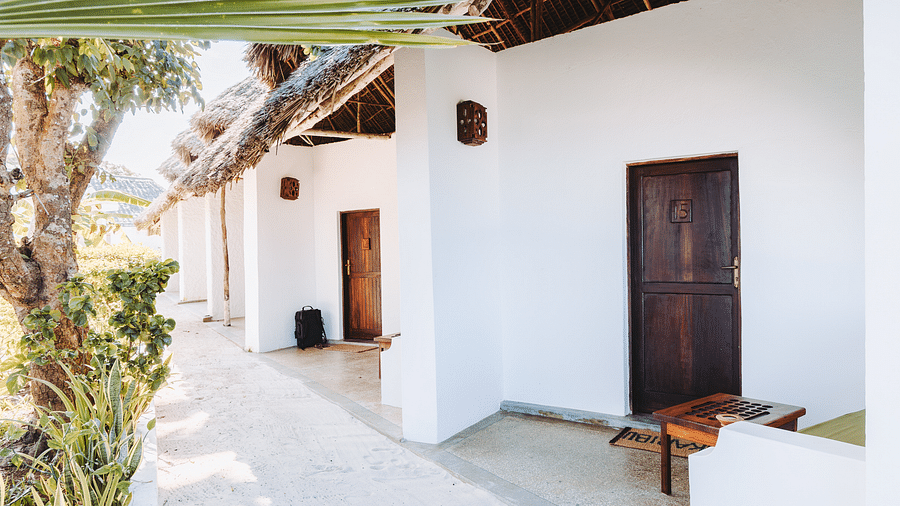 Exterior view of a row of buildings with thatched roofs and wooden doors, along a paved walkway with plants on the left - Shooting star Boutique Hotel.