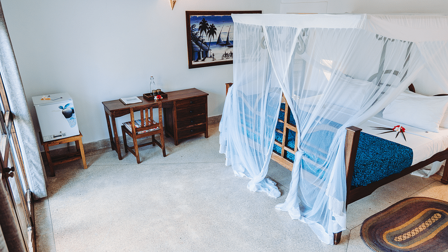 Interior view of a bedroom with a wooden four-poster bed with a mosquito net, a bedspread, a desk with a chair, a framed picture on the wall, and a small refrigerator - Shooting Star Boutique Hotel.
