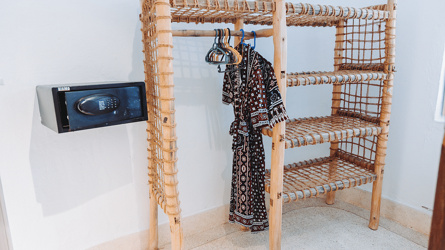 A wicker shelf unit with three shelves and a hanging dress, alongside a safe mounted on the wall - Shooting Star Boutique Hotel.