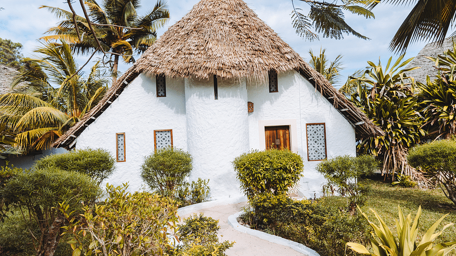 White house with a thatched roof surrounded by lush greenery and palm trees. A curved pathway leads to the wooden front door. Bright and sunny day with a clear blue sky - Shooting Star Boutique Hotel.