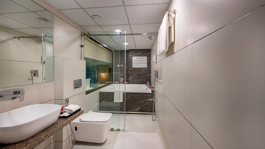 A modern hotel bathroom in Bengaluru features a sleek white sink on a stone countertop, a toilet, and a glass-enclosed bathtub - SK Lords Inn Ahmedabad