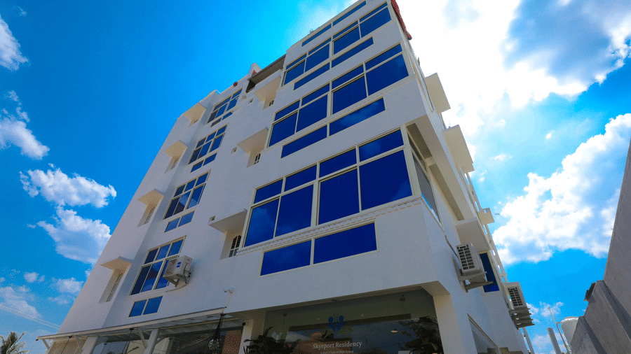 A low-angle shot of a multi-storey white building with large blue-tinted windows against a blue sky with clouds | Skyeport Residency
