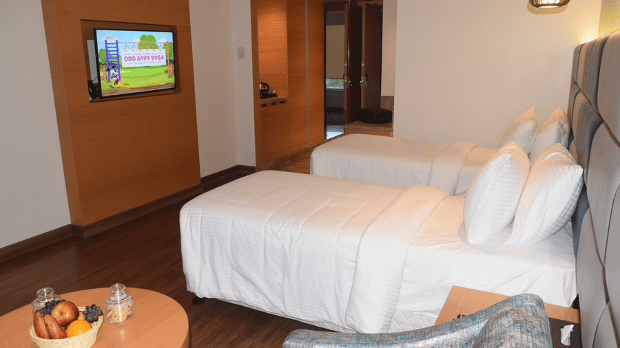 A standard twin room at SRM Hotel, Chennai with neatly made twin beds, a wall-mounted TV, wooden flooring, a small round table with a fruit bowl, and a cushioned chair next to the beds.
