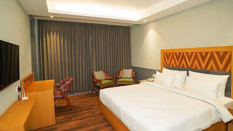 A brightly lit hotel room showing a neatly made bed with white linens and a view towards the window with drawn curtains| Deluxe Room | Sukama Resort & Spa, Nainital
