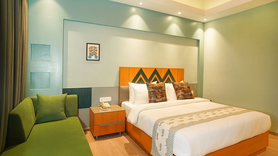 A hotel room showcases a neatly made bed with a green throw blanket, a bedside table with a lamp, and a green sofa | Deluxe Room with Valley View | Sukama Resort & Spa, Nainital