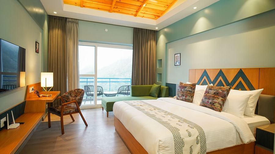 A spacious hotel room includes a large bed, a comfortable seating area with a sofa, and a balcony view of the hills | Deluxe Room with Valley View | Sukama Resort & Spa, Nainital
