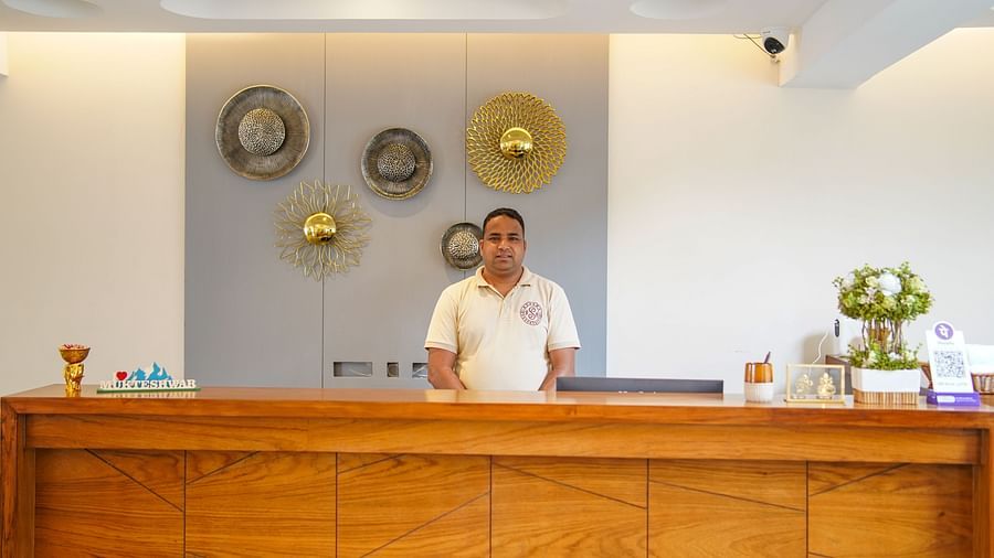 A hotel reception desk with a person standing behind it, and decorative plates on the wall behind them | Sukama Resort & Spa, Nainital