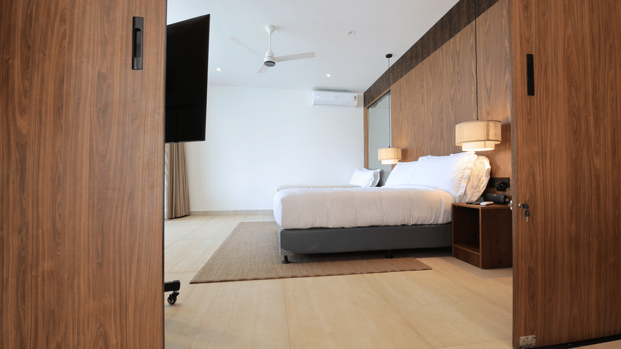A bedroom at SUKAVI featuring a king size bed, a wood-panelled accent wall, and a partially open sliding wooden door viewed from the entrance.