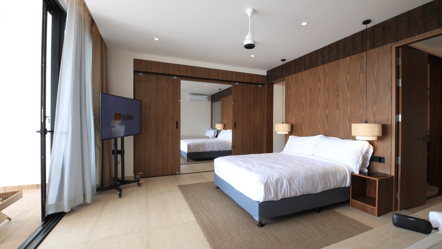 A bedroom at SUKAVI featuring a king size bed, a wall-mounted television, a ceiling fan, and large sliding glass doors that open onto a private balcony.