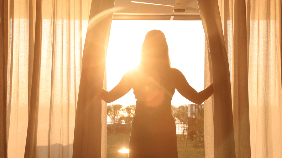 Silhouette of a person standing between open curtains, looking out through a sunlit window at SUKAVI, with natural light filling the room.