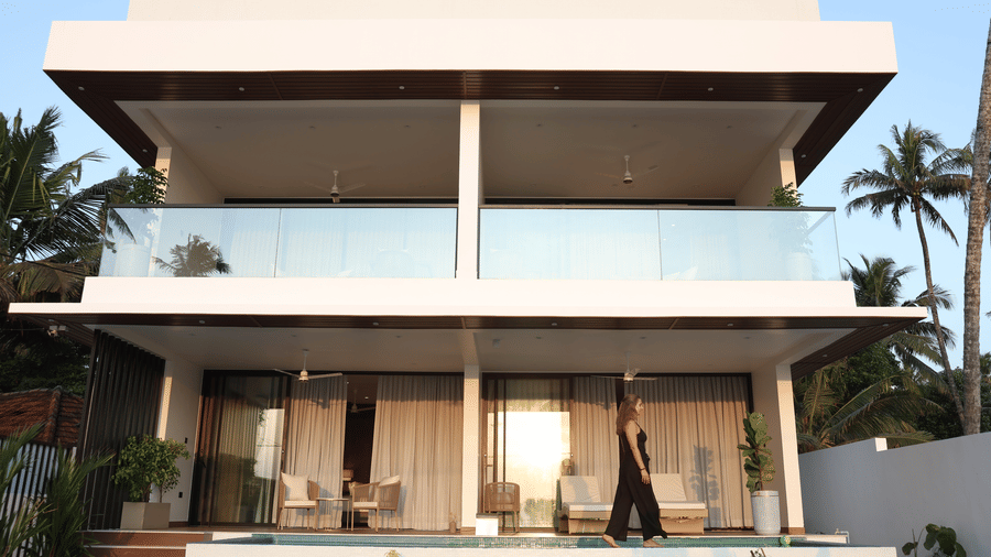 A multi-story modern building at SUKAVI, with balconies and glass railings, viewed from the front with a narrow pool and palm trees visible.
