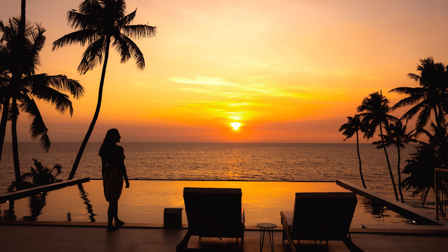 Silhouettes of a person standing and two lounge chairs beside an infinity pool at sunset at SUKAVI, surrounded by palm trees and overlooking the horizon.