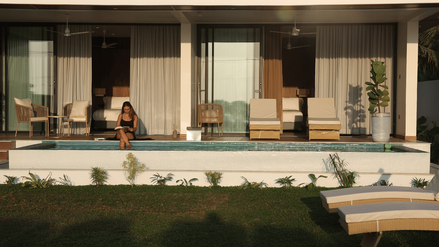 A person sitting at the edge of a small pool outside a modern ground-floor villa with sliding glass doors and outdoor seating at SUKAVI.