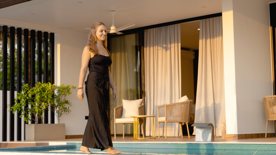 A person walks along the edge of an outdoor swimming pool in front of a patio area at hotel SUKAVI.