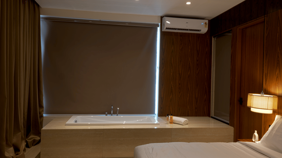Hotel room at SUKAVI with a bed, a window with closed blind, an air conditioning unit, a bedside lamp, and a bathtub visible in the background.