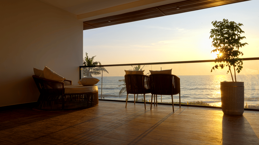 Balcony at SUKAVI overlooking the ocean at sunset with wooden flooring, a glass railing, 2 chairs, a lounge chair, and potted plants.