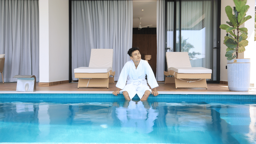 A person wearing a robe sits by the swimming pool with their feet in the water at SUKAVI, relaxing in front of patio lounge chairs.