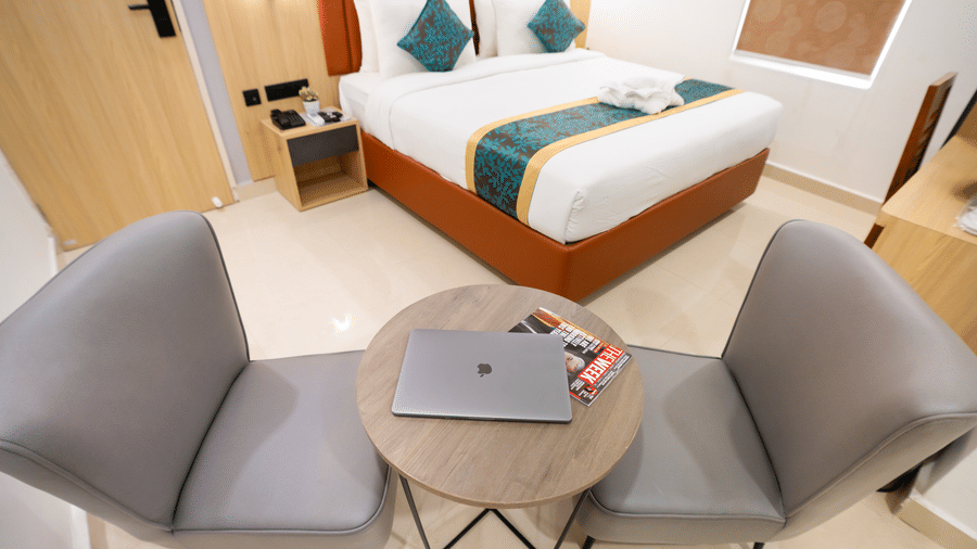 A close-up view of a small round table with a laptop and chairs on either side, with a large bed in the background at Sunrise Business Hotel, Hyderabad.
