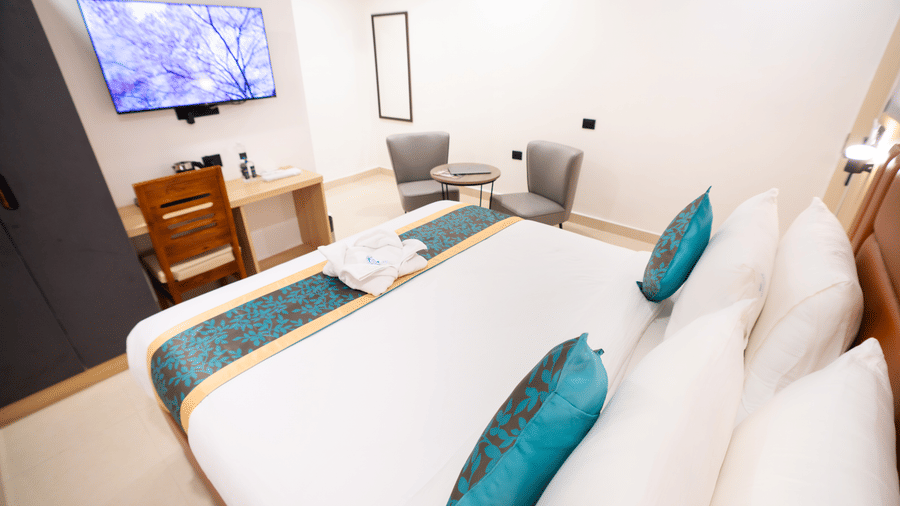 View of the room from beside the headboard, showing a neatly made bed facing a wall-mounted TV and a study table with a chair below at Sunrise Business Hotel, Hyderabad.