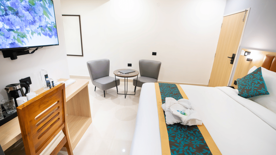 A view of the room with a large TV, a small table with chairs to the side, wooden interiors, and a spacious bed at Sunrise Business Hotel, Hyderabad.