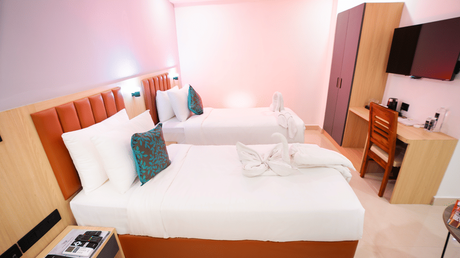 Side view of a spacious room with twin beds, a bedside table, and a wall-mounted television at Sunrise Business Hotel, Hyderabad.