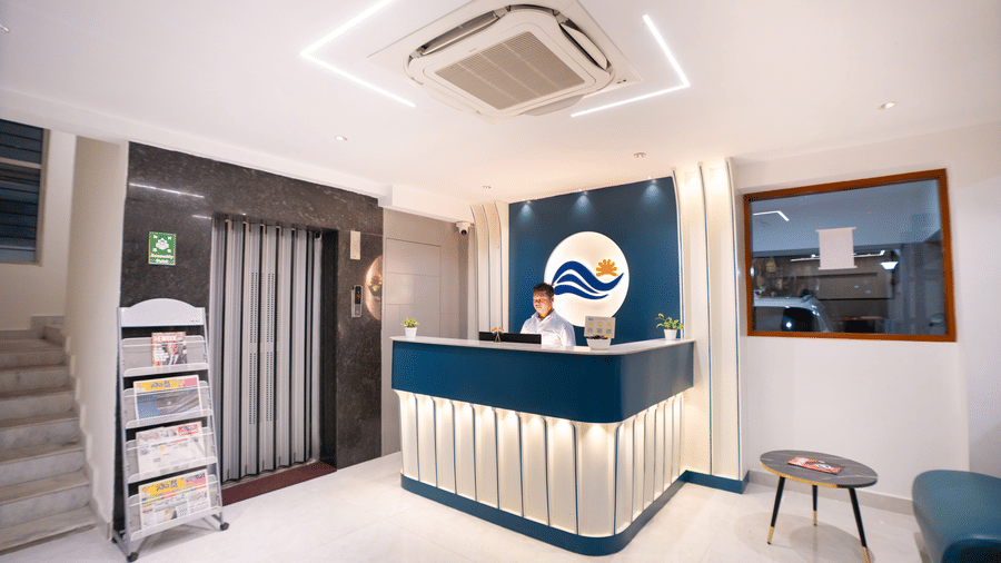 A diagonal view of the reception area with a large desk and a staff member standing behind it, a seating area on one side, and a newspaper stand on the other at Sunrise Business Hotel, Hyderabad.