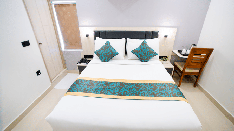View of a room with a spacious bed, bedside table, wall-mounted A/C above the headboard, and a closed door to the side at Sunrise Business Hotel, Hyderabad.