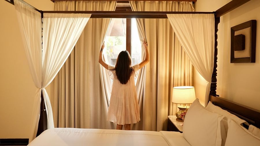 A bedroom with a lady opening the curtains covering the window for a view outside.