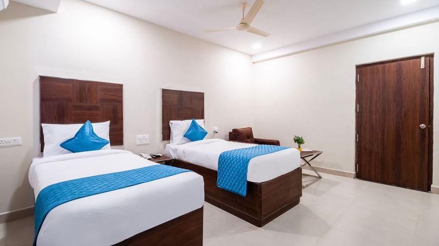 Twin beds with blue accents placed in a spacious bright room with wooden furniture and ceiling fan at Swarna's The Capital.