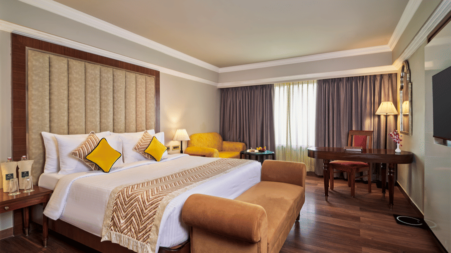 The Club Room at Swosti Premium, Bhubaneswar, featuring double bed, bedspreads, pillows, chairs, tables, cushions, a television, and large windows.