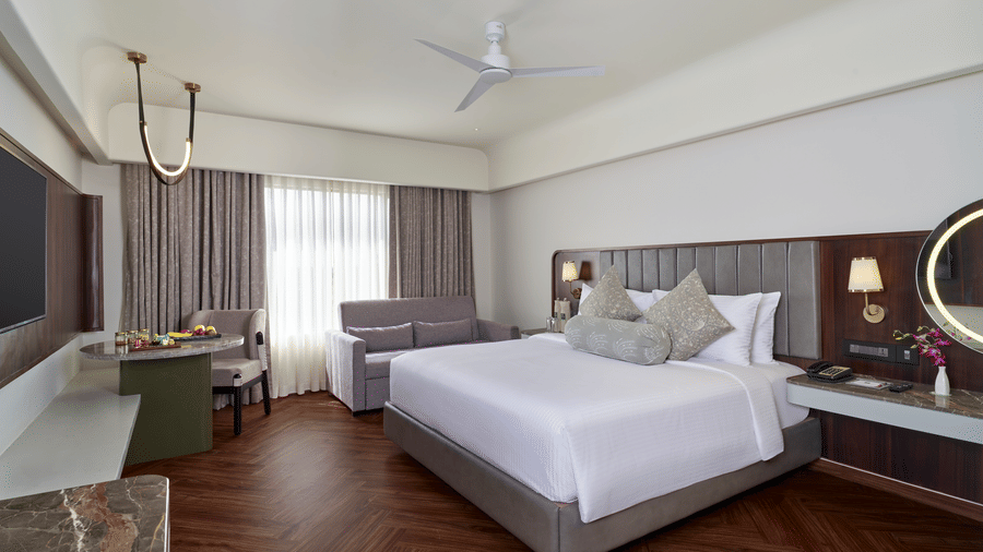 The Elite Room at Swosti Premium, Bhubaneswar, featuring a double bed, bedspreads, pillows,  cushions, a table, large windows covered in curtains, a mirror, a television,  wall mounted lamps and a fan. 