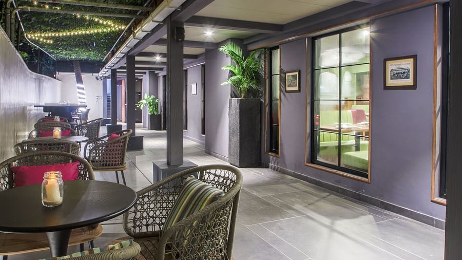 A dimly lit lounge area with several round tables and chairs, as well as a long   | T24 Retronch against the wall. The space is decorated with plants, artwork, and warm lighting, creating a cosy atmosphere.