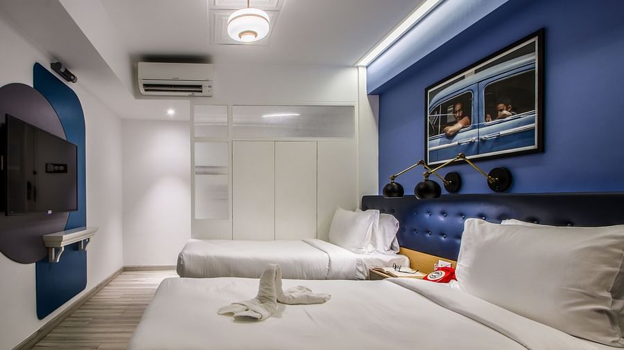A spacious and well-lit hotel room with twin beds, a TV mounted on the wall, and a large mirror  | T24 Retro