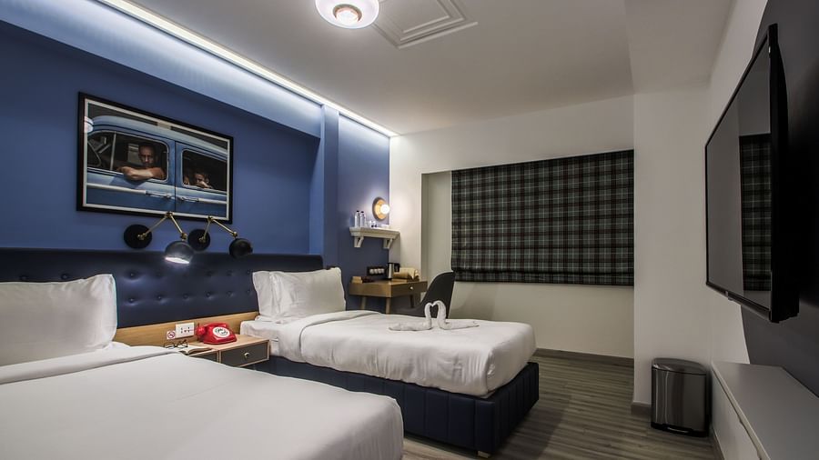A well-lit hotel room with twin beds, a desk, a TV, and a large window. The walls are painted a deep blue, and there is a framed picture above the bed   | T24 Retro