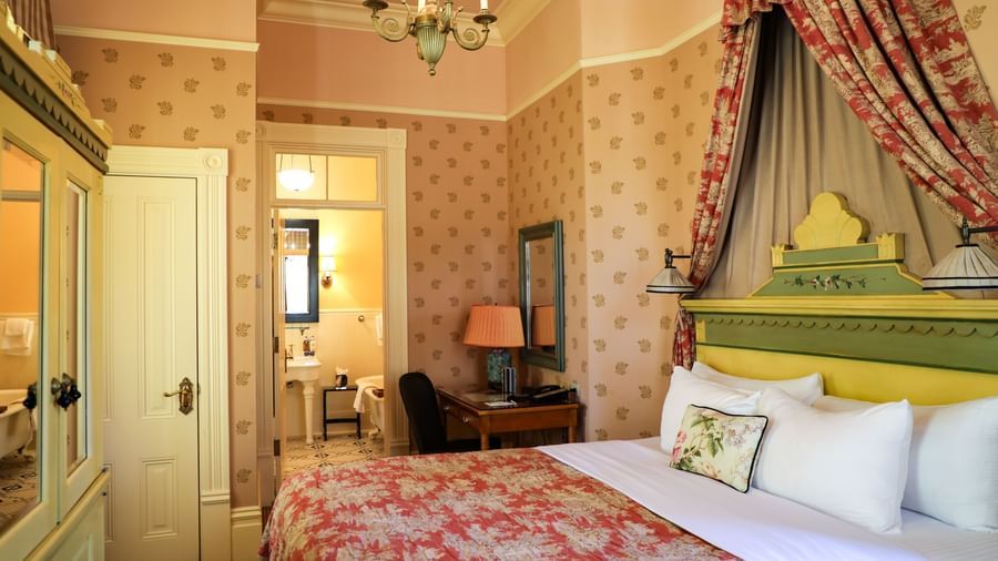 A bedroom at the Tallman Hotel contains a bed with a canopy, a dresser, and an open door to a bathroom.