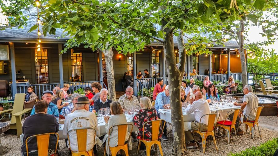 A crowd seated at outdoor dining arrangement and eating under tree shade at Tallman Hotel.