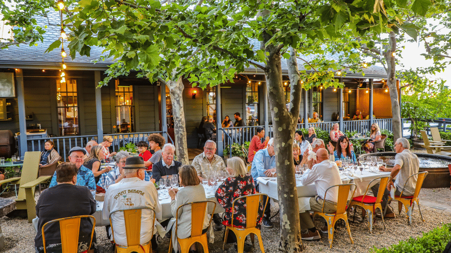 A crowd seated at outdoor dining arrangement and eating under tree shade at Tallman Hotel