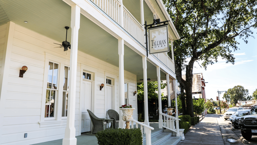 Side view of the Tallman Hotel shows an inviting porch, white facade, and a sign hanging over the entrance.