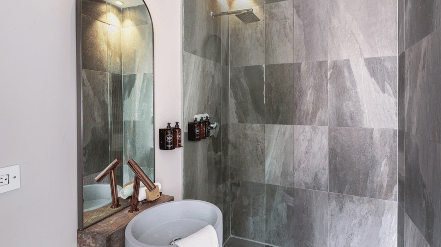 A bathroom with a glass-enclosed walk-in shower, stone-effect walls, an arched mirror, and bathroom fixtures in Suite 35 at Tamarind Hills Resort and Villas.