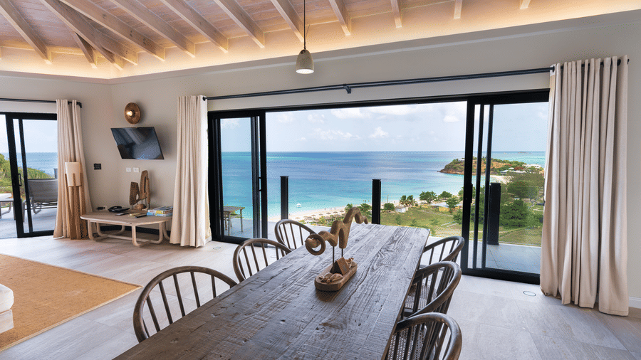 A dining area with a wooden table and chairs beside large sliding glass doors overlooking the swimming pool, coastline, and sea at Suite 35, Tamarind Hills Resort and Villas.