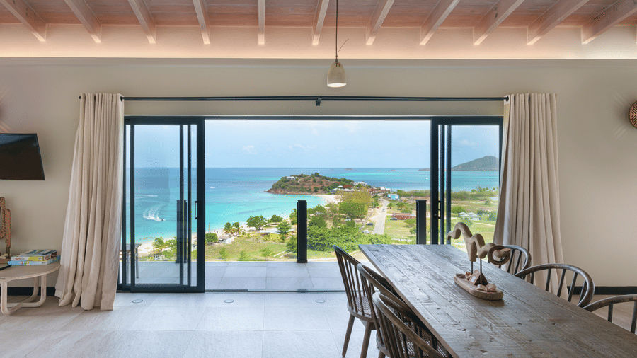 A dining area with a long table and chairs positioned beside large glass doors overlooking the swimming pool, sea, and coastline at Suite 35, Tamarind Hills Resort and Villas.