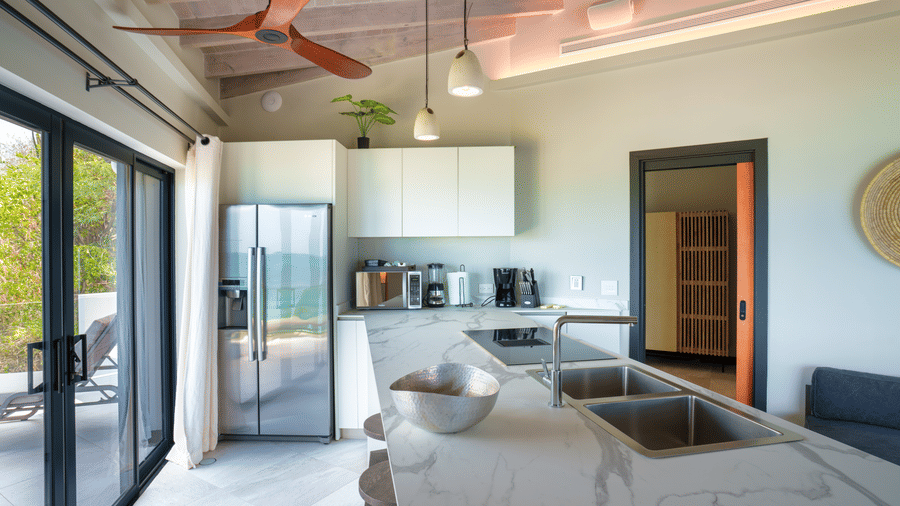 A living and kitchen area with a sofa, kitchen counter, dining space, ceiling fan, and large glass doors opening outdoors in Suite 35 at Tamarind Hills Resort and Villas.